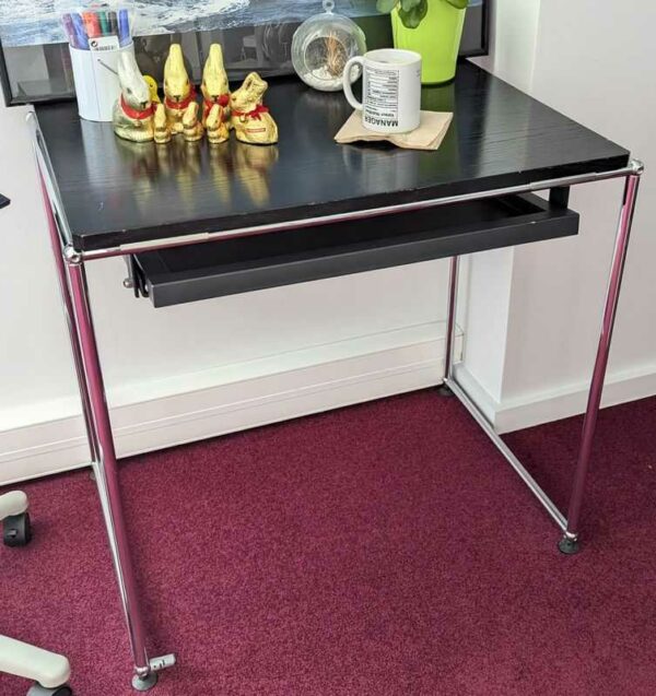 Table de marque USM HALLER, plateau en bois de couleur noire avec une tablette rétractable en ceinture, reposant sur une structure en acier chromé. 70 x 76 x 54 cm. Localisation : Canada 1er (2B01)