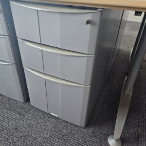 Bureau d'angle en bois couleur miel clair reposant sur un piètement métallique triple en acier gris. Dimensions: 160 x 160 cm. On y joint son caisson à hauteur d'appui. Quantités disponibles: 830 unités. Localisation : 8 av. de Lunca - 78180 Montigny-le-Bretonneux. – Image 3